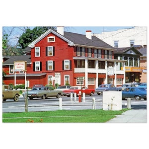 May include: A vintage photograph of a street scene featuring a red building with black shutters and a white trim. Classic cars are parked on the street. A sign indicates "Central Markets" and "Parking".