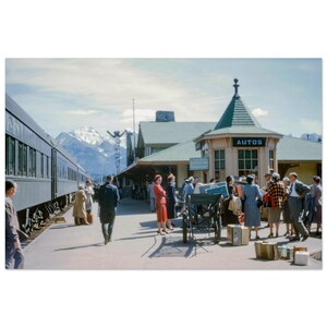 May include: Vintage photograph of a train station with a train on the left and a building with "AUTOS" on the right. People are gathered on the platform, with luggage and a horse-drawn carriage. Snow-capped mountains are visible in the background.