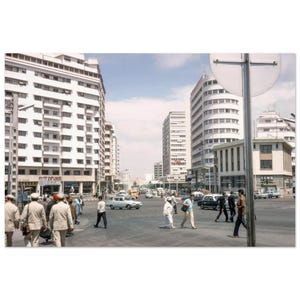 Pode incluir: Fotografia vintage de uma cena de rua movimentada com pedestres atravessando uma rua larga. Edifícios altos e brancos ladeiam a rua sob um céu azul. Carros e ônibus são visíveis, e uma placa de rua está em primeiro plano.