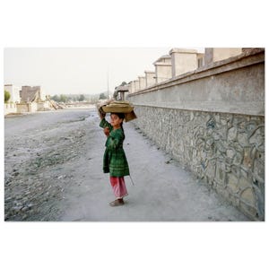 May include: A young person in a green dress and pink pants carries a tray on their head while walking on a dirt path. A stone wall and buildings are in the background. The image is taken in daylight.