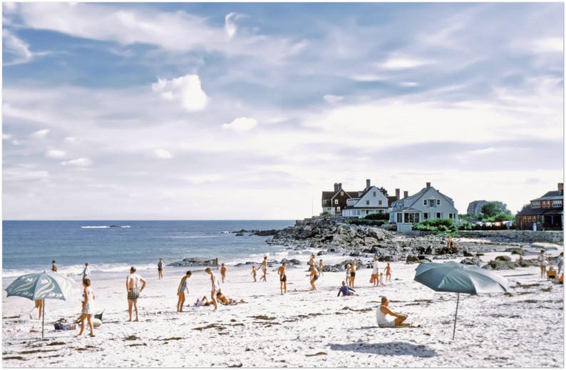 May include: A vintage photograph of a beach scene with people enjoying a sunny day. The image shows a sandy beach, blue ocean, and a cloudy sky. Several people are on the beach, some with umbrellas.