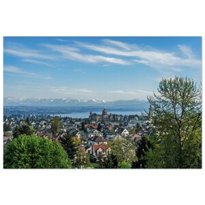 May include: A panoramic view of a town featuring numerous houses with pitched roofs, surrounded by lush green trees. In the distance, a large building with a prominent dome stands near a body of water, backed by snow-capped mountain peaks under a blue sky.