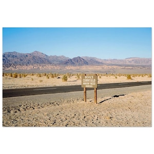 May include: A rectangular brown sign with white text reading "DEVIL'S CORNFIELD ARROW-WEED SHOCKS" mounted on two wooden posts. The sign stands in a dry, sandy desert landscape next to a paved asphalt road, with rugged mountains in the distance.