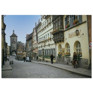 Könnte beinhalten: Eine Kopfsteinpflasterstraße in einer europäischen Stadt mit bunten Gebäuden und Blumenkästen. Ein Uhrturm ist im Hintergrund zu sehen. Ein Oldtimer und Fußgänger sind auf der Straße unterwegs.