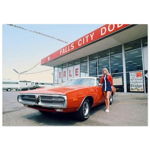 Könnte beinhalten: Ein orangefarbener Dodge Charger mit weißem Dach steht vor einem Falls City Dodge Autohaus. Das Wort "SALE" ist auf dem Gebäude zu sehen. Eine Frau in einem blau-roten Kleid steht neben dem Auto.