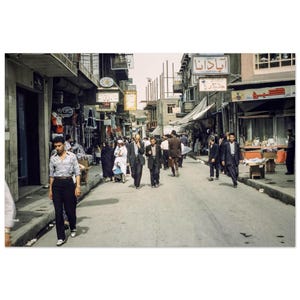 May include: A vintage photograph captures a bustling street scene. People walk along a paved road lined with shops and buildings. Signs in a foreign language hang above the shops. The image has a warm, nostalgic tone, suggesting a historical or cultural setting.