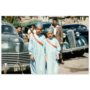 May include: Vintage photograph featuring two young boys in light blue robes and decorated hats, standing in front of classic cars. The boys wear red and white sashes. Other people are visible near the cars. The image has a nostalgic feel.