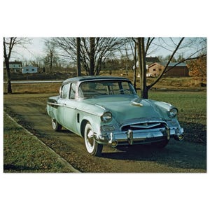 Könnte beinhalten: Eine hellminzgrüne Oldtimer-Limousine mit Chromstoßstangen, runden Scheinwerfern und Weißwandreifen. Das Fahrzeug verfügt über eine markante Kühlerfigur, eine Panorama-Windschutzscheibe und eine schlanke Karosserie auf einem Kiesweg.
