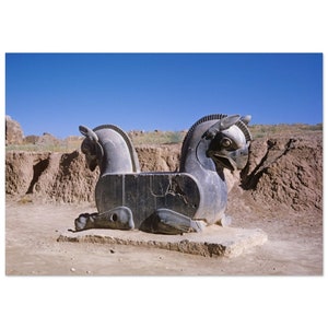 May include: A large, gray stone sculpture of a mythical creature with two heads, resembling a winged horse, sits on a dirt mound. The sculpture is in a desert setting with a blue sky in the background.