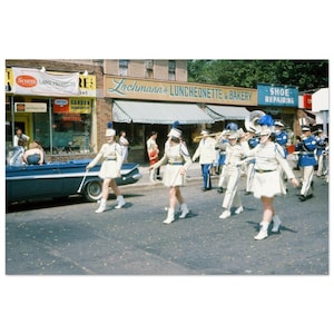 Puede incluir: Una fotografía vintage de un desfile de banda de música. Los miembros de la banda visten uniformes color crema con detalles azules y dorados. La imagen muestra un coche clásico y escaparates, incluyendo una panadería y una zapatería.