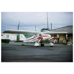 May include: A white and red twin-engine airplane parked on an asphalt surface. The plane has two propellers and the text "LIMBE AIR TRANSPORT" and "TJ-ACB" on the side. The background includes a building and a cloudy sky.