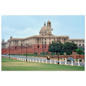 May include: A large, beige-colored building with a domed roof and multiple columns. A road with a white fence runs in front of the building, with green grass on either side. The Indian flag flies from a tower.