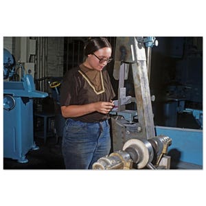 Può includere: Una persona che indossa una maglietta marrone a maniche corte con colletto beige e jeans blu utilizza una levigatrice a nastro verticale in un'officina. L'attrezzatura industriale presenta un telaio in metallo e un nastro abrasivo rotante.