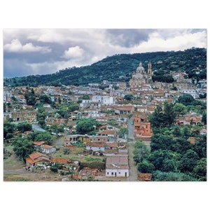 May include: An aerial view of a small town nestled in a valley, with a large white church with a dome at the top of the hill. The town is surrounded by lush green trees and hills.