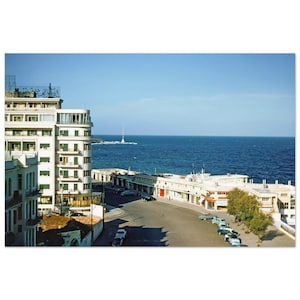 May include: A vintage photograph captures a coastal city scene under a clear blue sky. White buildings line the street, with parked cars and the sea in the background. A lighthouse stands in the distance, adding to the seaside ambiance.