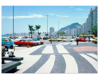 Può includere: Fotografia vintage di una città balneare in una giornata di sole. Un attraversamento pedonale conduce alla spiaggia, con auto e persone. Palme ed edifici sullo sfondo sotto un cielo azzurro. L'immagine ha un'estetica retrò.