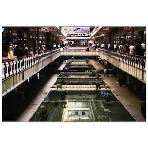 May include: An interior shot of a multi-level shopping mall with walkways, water features, and shops. People are seen walking on the upper levels and along the water features. The architecture features a mix of concrete, metal, and glass, with neon signs.