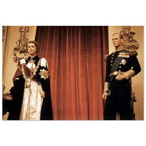 May include: A colour photograph of a man and woman in formal attire standing in front of a red velvet curtain. The woman is wearing a black and white gown with a gold chain and a red sash. The man is wearing a black military uniform with gold trim and a sword.