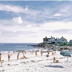 May include: A vintage photograph captures a sunny beach scene with people enjoying the day. The sky is blue with scattered clouds. Beach umbrellas provide shade. Houses are visible on a rocky outcrop in the background.