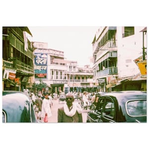 May include: Vintage street scene featuring a man in a suit and scarf, with buildings and cars. A sign in a foreign language is visible. The image has a retro aesthetic, with a focus on the architecture and people.