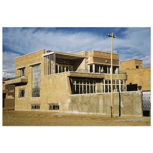 May include: A multi-story tan brick building featuring a flat roof, a second-floor balcony with a metal railing, and large rectangular windows. A tall wooden utility pole stands in front of the structure, which is set against a blue sky.