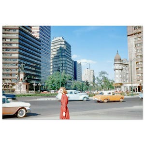 May include: A retro photograph depicts a city street with high-rise buildings and vintage cars. A person in a red dress is on a pedestrian crossing. The scene is illuminated by sunlight, under a clear blue sky.