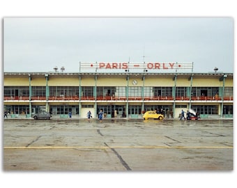 Op de afbeelding: Een vintage foto van het terminalgebouw van de luchthaven Paris-Orly. Het gebouw is een verdieping hoog met een plat dak en een groot bord met de tekst "PARIS - ORLY" in rode letters. Er lopen verschillende mensen voor het gebouw en een gele auto staat geparkeerd op de voorgrond.