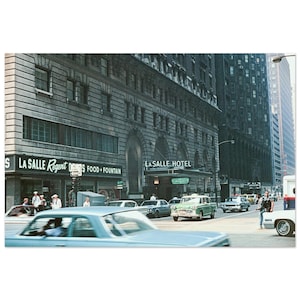 May include: A vintage street scene featuring the La Salle Hotel and La Salle Regent building with a food and fountain sign. Several mid-century automobiles are driving on the city street, including a light blue car in the foreground.
