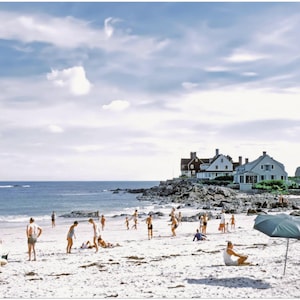 May include: A vintage photograph captures a sunny beach scene with people enjoying the day. The sky is blue with fluffy clouds. Beach umbrellas provide shade. Houses are visible in the background, and the ocean is calm.