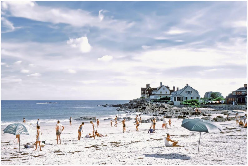 May include: A vintage photograph captures a sunny beach scene with people enjoying the day. The sky is blue with fluffy clouds. Beach umbrellas provide shade. Houses are visible in the background, and the ocean is calm.
