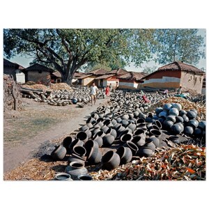 May include: A large group of black clay pots are arranged in rows on a dirt path in front of a village. The pots are all different sizes and shapes, and some are stacked on top of each other. There are several people walking in the background.