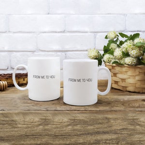 Peut inclure: Deux tasses en céramique blanche avec du texte noir qui dit "De moi à toi". Les tasses sont sur une surface en bois avec un panier en osier de fleurs blanches en arrière-plan.