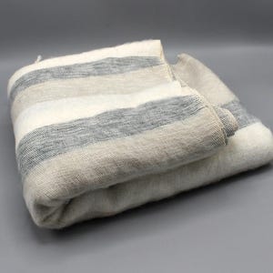 May include: Folded scarf with horizontal stripes in shades of gray, cream, and beige. The scarf appears to be made of a soft, woven material, with a slightly textured surface. The neutral color palette suggests a versatile accessory.
