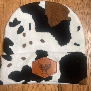 Op de afbeelding: Witte, zwarte en bruine gebreide beanie met een koe-printpatroon. Een bruin leren patch met een zwart-witte illustratie van een koeienkop is op de voorkant van de beanie genaaid.