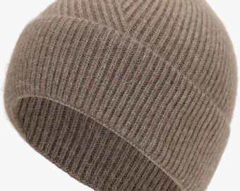 May include: A taupe beanie hat featuring a vertical ribbed knit pattern. The hat has a folded cuff at the bottom edge and a rounded crown construction with converging knit lines that meet at the top center, creating a textured appearance.