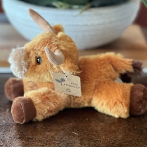 May include: A plush brown and white Highland cow stuffed animal with a tag that says "Eco Friendly".