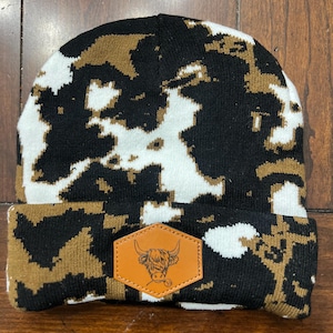 Könnte beinhalten: Eine schwarze, weiße und braune Strickmütze mit einem braunen Lederpatch, auf dem eine Comic-Highland-Kuh abgebildet ist. Der Patch hat eine sechseckige Form.