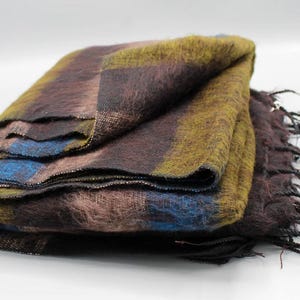 Pode incluir: Um cobertor retangular dobrado com um padrão xadrez. O cobertor apresenta uma mistura de cores, incluindo marrom, azul e amarelo. As bordas têm franjas. O cobertor é feito de um material macio e difuso.