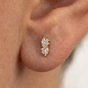 May include: A gold stud earring featuring a cluster of clear, faceted stones in a vertical arrangement. The design includes two marquise-cut stones at the top and bottom, accented by three smaller round-cut stones positioned in the center of the piece.