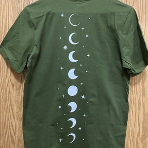 May include: A green t-shirt with a white moon phases design on the back. The design features a series of moons in different phases, from a crescent to a full moon, with stars scattered around them.