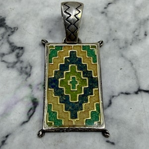 May include: Silver pendant with a rectangular, geometric design in shades of green, blue, and gold. The pendant has a zig-zag patterned bail and small decorative accents at each corner. The design is reminiscent of Southwestern or Native American art.