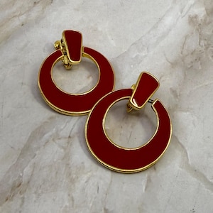 Puede incluir: Un par de pendientes de aro rojos y dorados. Los pendientes tienen un diseño circular con un acento rectangular en la parte superior. Los aros son rojos con un borde dorado y un cierre de clip dorado.