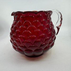May include: A red glass pitcher with a clear glass handle. The pitcher has a textured, scalloped rim and a textured,  scalloped body.