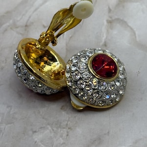 Könnte beinhalten: Ein Paar goldfarbene Ohrclips mit einem roten Strassstein in der Mitte und klaren Strasssteinen, die die Mitte umgeben. Die Ohrringe haben eine weiße Rückseite.