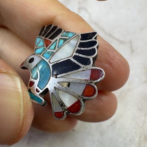 May include: A silver-toned bird-shaped pin with inlaid turquoise, black, white, and red stones. The bird has a detailed design with a stylized wing and head, showcasing a Native American-inspired aesthetic. The pin is held in a hand.