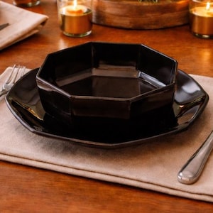 May include: A dining table set for a meal. The table is set with black plates and bowls, silverware, and wine glasses. A floral centerpiece and candles add to the ambiance. The table is made of wood.