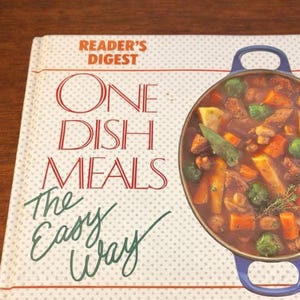 May include: A cookbook titled "One Dish Meals The Easy Way" by Reader's Digest. The cover features a colorful illustration of a stew in a blue pot. The book is on a wooden surface.