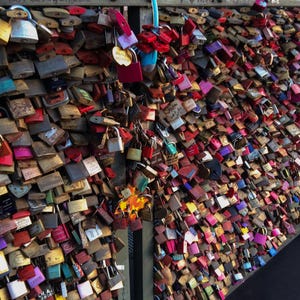 Può includere: Un muro ricoperto da numerosi lucchetti di vari colori, tra cui oro, rosso, rosa e blu. I lucchetti sono attaccati a una struttura metallica, creando una superficie densa e strutturata. Alcuni lucchetti hanno piccole etichette.