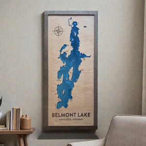 May include: A rectangular wooden wall map of Belmont Lake in Havelock, Ontario, featuring a deep blue laser-cut lake depth design on a light wood background, enclosed in a dark wood frame with a compass rose in the upper left corner.