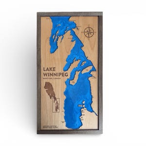 May include: A framed wooden map of Lake Winnipeg, Manitoba, Canada. The lake is represented in blue resin, set against a natural wood background. The text "Lake Winnipeg" is visible, along with a compass rose.